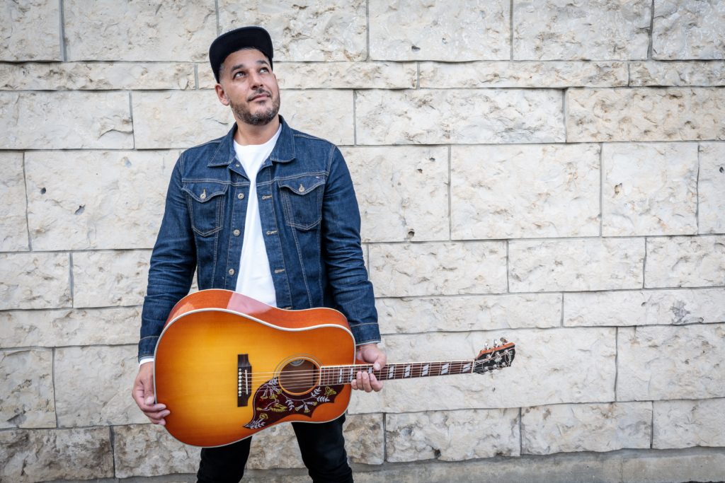 Adrian Sutherland standing in front of a brick wall holding a guitar. 